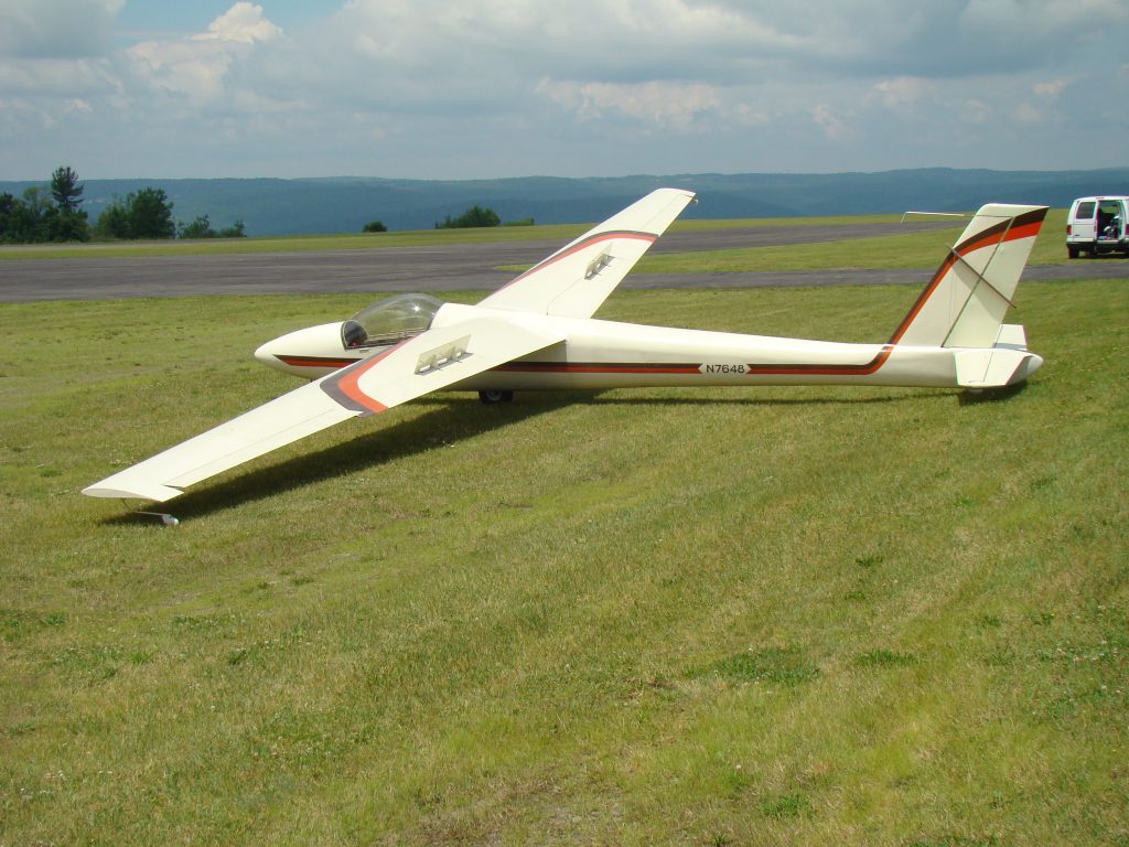 A cream colored Schweizer 1-34 glider sits in a grass field near a runway in the background. The glider has orange and black detail stripes on the wings and fuselage.
