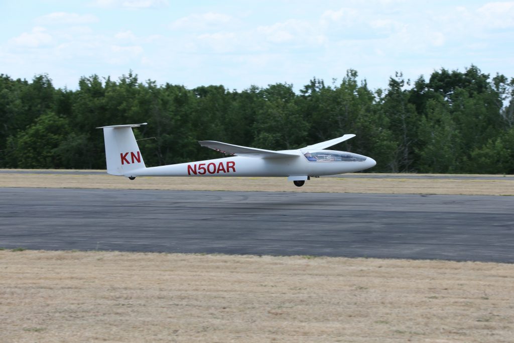 A white Glasflugel glider approaches touchdown on a runway (heading from left to right) with green trees in the background.