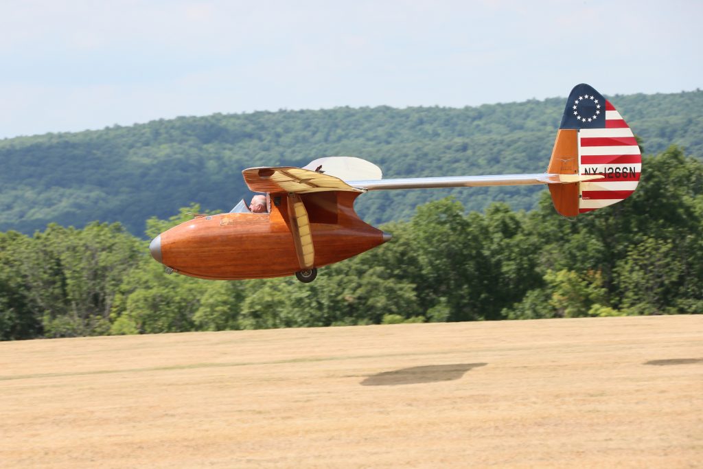 A wooden Bowlus Baby Albatross glider flying from right to left approaching to land on a brown field with trees in the background.