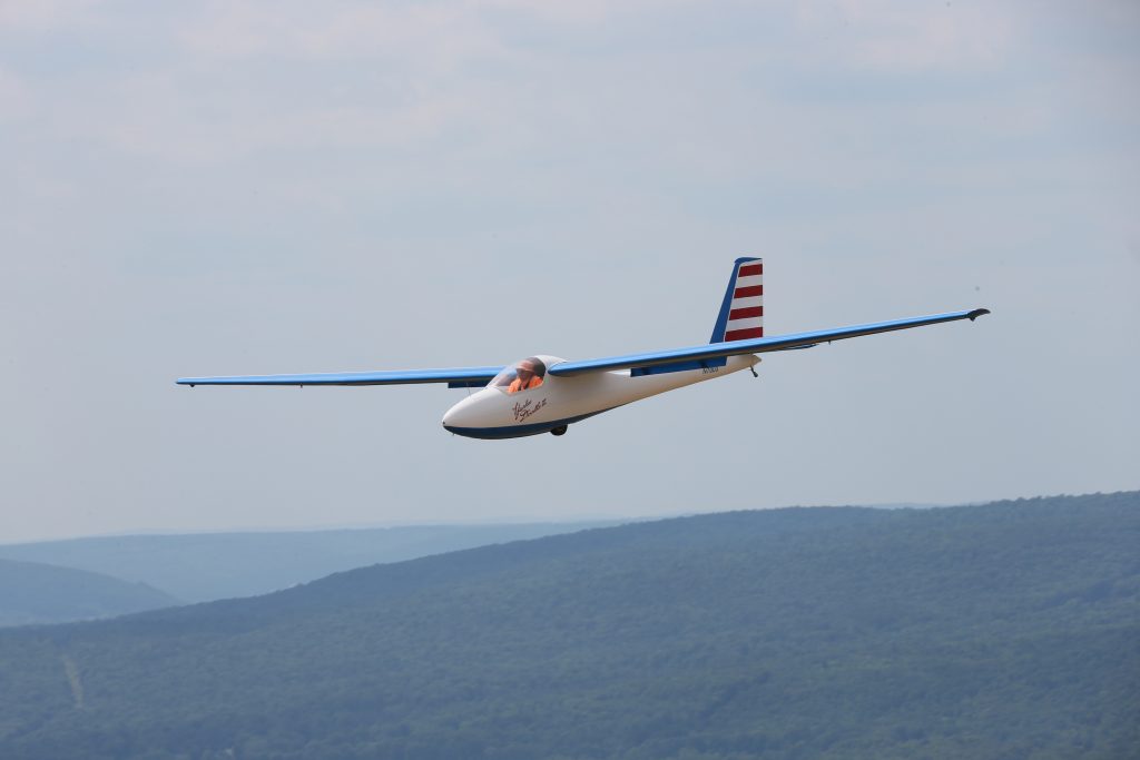 A single-seat Laister glider flies (right to left) over hills in a gray sky. The glider is has a white body, blue wings and red and white stripes on the tail.